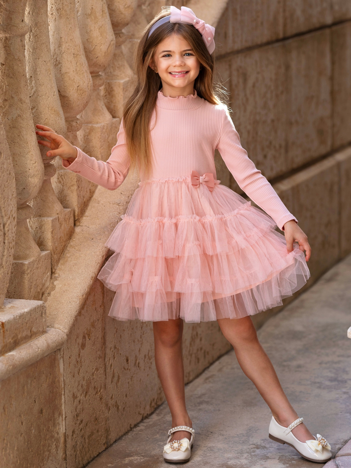Girl wearing a blush pink ribbed knit dress with a tiered tulle skirt and bow detail for birthdays and special occasions.